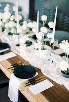 Black plate stands next to gold cutlery on a festive table in front of a glass and bouquets of white flowers photo