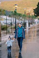 sonriente madre con un pequeño niña caminando participación manos a lo largo un mojado calle con brillante linternas foto