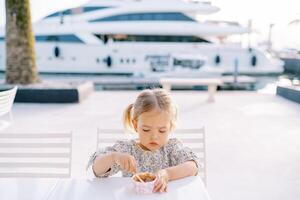 Little girl picks ice cream with a spoon, holding a glass with her hand on a table on the seashore photo