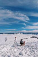 Mom with a small child sit on a sled near a snowman and look at the snow plain. Back view photo