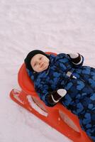 Little boy in overalls and mittens lies on a sled in the snow photo