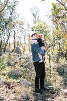 Little girl hugs her dad by the neck, standing in a green park, sitting in his arms. Side view photo