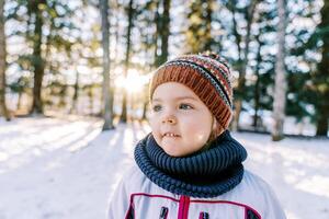 pequeño sonriente niña en un sombrero y bufanda soportes en un Nevado bosque y mira lejos foto