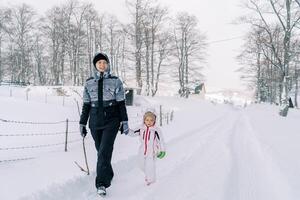 Mother and a little girl walk holding hands along a snowy road on the edge of a forest photo