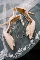 Wedding rings lie on an embroidered veil near the bride golden high-heeled shoes photo