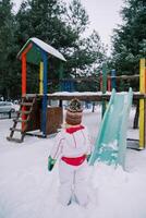 Little girl stands near a snow-covered colorful slide and looks at it. Back view photo