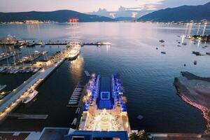 Laid tables on the pier near a festively lit stage in front of a long seaside pool. Drone photo