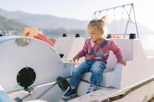 pequeño niña se sienta en el asiento de un catamarán y mira a el pedales foto