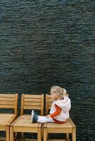 Little girl sits on a row of wooden chairs against the wall and looks at her shoes. Side view photo