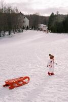 Little girl is walking along a snowy hill, dragging a sled on a rope behind her. Side view photo