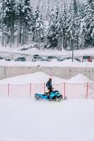 Man in a helmet riding a snowmobile up the mountain along a red fence while standing photo