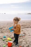 Little girl with a toy bucket in her hands stands on a sandy beach surrounded by colorful toys photo