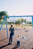 mamá se sienta en un cadena columpio en el patio de recreo y mira a un pequeño niña en pie siguiente a su foto