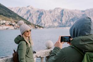 Husband photographs his wife leaning on the railings on the seashore overlooking the mountains. Back view photo