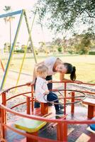 Mom leaned over to a little girl sitting on a carousel on the playground photo