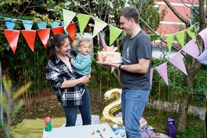 Little girl sits in her mother arms and looks at a colorful cake in her dad's hands in the garden photo