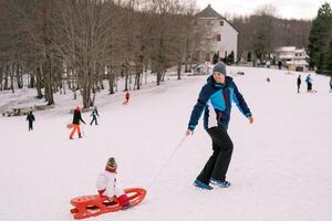 Smiling dad climbs a hill with a small child in a sled, looking back. Side view photo