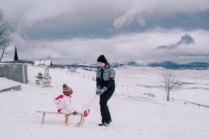 Smiling mother carries little girl on wooden sled along snowy hill photo