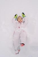 pequeño niña mentiras en un ventisquero haciendo un nieve ángel con su ojos cerrado foto
