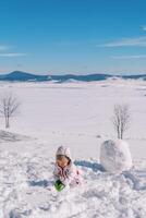 pequeño niña hace un grande bola de nieve para un monigote de nieve en pie en un Nevado claro foto