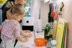 pequeño niña Ayudar su madre amasar Pizza masa a el cocina mesa foto