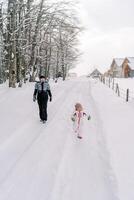 madre y un pequeño niña caminar a lo largo un Nevado la carretera en un pueblo a el borde de el bosque foto