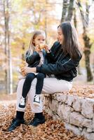 pequeño niña roe un manzana mientras sentado en su madre brazos en el bordillo en un otoño parque foto