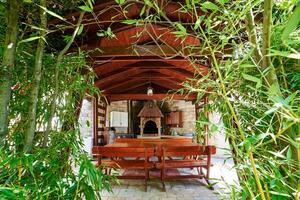 View through the green branches of the garden to a wooden gazebo with a stove and kitchen cabinets photo