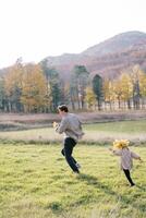 Little girl in a wreath of autumn leaves runs after her dad on the sunny lawn photo