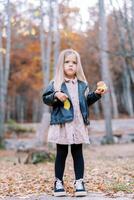 pequeño niña con un manzana en uno mano y un amarillo hoja en el otro es en pie en un otoño parque foto