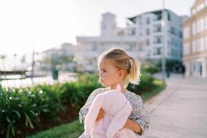 Little girl with a pink plush rabbit in her hands walks along the yard of a multi-story building and looks to the side photo