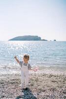 Little girl with a toy walks along a pebble beach, balancing with her hand photo