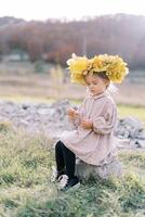 Little girl in a wreath of autumn leaves sits on a stone on a green lawn and examines a blade of grass in her hands photo