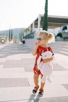 Little girl walks along a sunny embankment with a toy rabbit under her arm, picking her nose photo