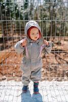 Little girl unzips her hood in overalls while standing near a metal fence photo