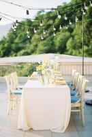 Long festive table with bouquets of flowers on the terrace with hanging garlands of light bulbs photo