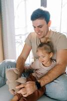 Dad plays with a little girl with a teddy bear sitting with her on the bed photo