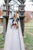 Little girl slides down a slide on a wooden playground holding the handrails photo