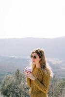 Smiling female traveler with a glass of coffee stands on a mountain photo