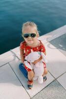 Little girl with a pink toy rabbit sits on a bollard on the pier and looks up. Top view photo