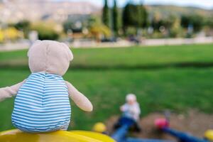 Teddy bear in a striped t-shirt sits on a swing in the playground. Back view photo