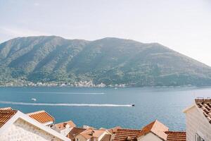 View over the red tiled roofs of houses to motor yachts sailing across the bay against the backdrop of mountains in a light haze photo