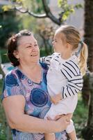 pequeño niña mirando a sonriente abuela mientras sentado en su brazos en el jardín foto