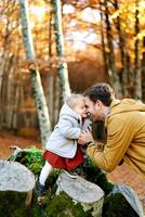 papá sostiene manos y toques su frente a el frente de un pequeño niña en pie en un tocón en un otoño parque foto