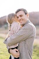 Little girl whispers in the ear of a smiling dad while sitting in his arms on a green meadow photo