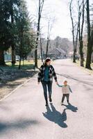 Mom and a little girl walk holding hands along an asphalt road in a sunny forest photo