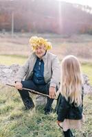 pequeño niña mira a su abuelo sentado en un Roca en el césped en un guirnalda de amarillo hojas. espalda ver foto