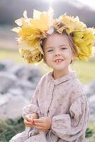 Little smiling girl in a wreath of autumn leaves sits on a stone on the lawn photo