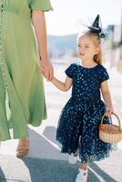 mamá Guías por el mano un pequeño niña en un vestir y un bruja sombrero con un cesta en su mano. recortado foto