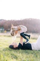 Smiling mother lifting little girl with arms outstretched to sides while lying on green lawn photo